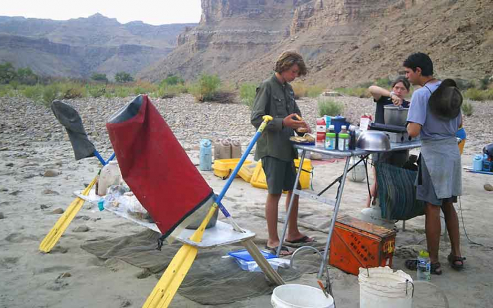Three people stand around a table set up in a desert landscape and prepare food.